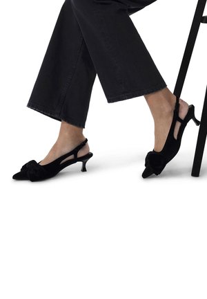Schwarze Samt-Slingback-Absätze mit einer spitzen Zehenpartie und einer Schleifendetail. Sie verfügen über einen niedrigen Stiletto-Absatz und werden mit schwarzen weiten Hosen kombiniert.