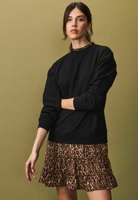 Hanorac negru cu guler texturat, asortat cu o fustă mini din leopard cu volane și straturi. Fundalul este de un verde stins.