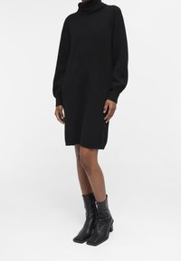 Schwarzes Strickkleid mit hohem Rollkragen, langen Ärmeln und lockerer Passform, das ein strukturiertes Muster aufweist und bis zur Mitte des Oberschenkels reicht. Schwarze Knöchelstiefel.