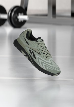 Chaussure de sport Reebok vert clair avec semelle noire et détails flottant au-dessus du sol de la salle de sport, haltère flou en arrière-plan.