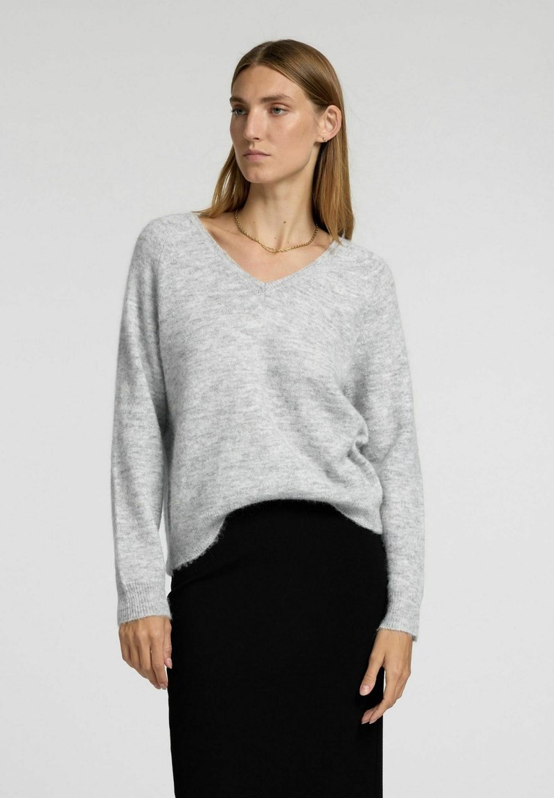 Grå strikket sweater med V-hals, lange ærmer og en afslappet pasform, parret med en sort nederdel. Blød tekstur og slightly længere bagkant.