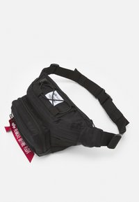 Mochila de cintura preta em nylon com um bolso principal com fecho zip, alça ajustável, cordas elásticas na parte de cima e uma etiqueta vermelha 'remover antes do voo'.