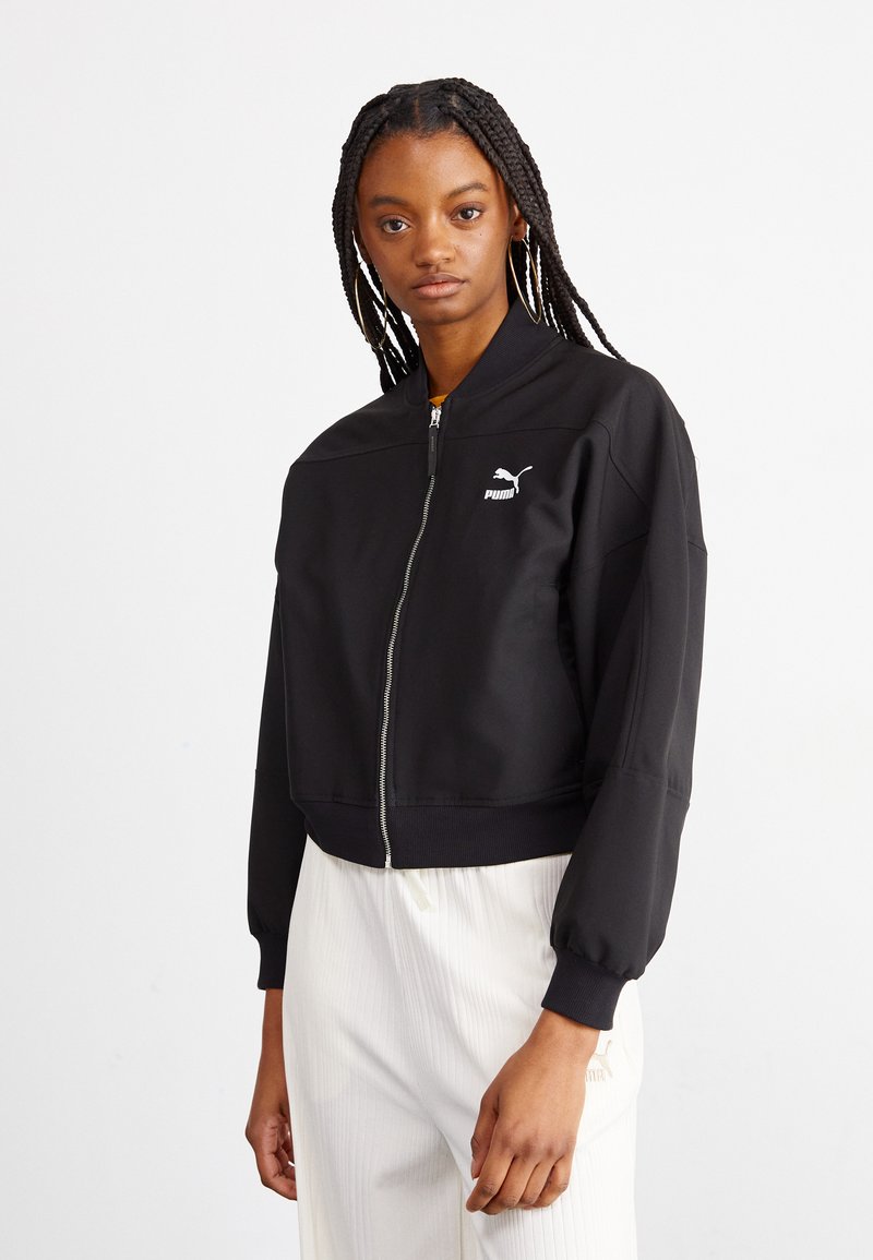 Veste bomber noire courte avec zip, poignets côtelés et logo Puma blanc sur la poitrine gauche. Tissu lisse avec une coupe décontractée.