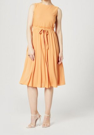 Femme portant une robe midi orange plissée sans manches avec une ceinture nouée à la taille et des sandales blanches à talons hauts sur un fond uni.
