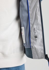 Kevyt takki sinisävyisessä ja harmaassa, verkkovuorella. Varustettu tuulenpitävyyttä ja vettä hylkivyyttä osoittavilla tarroilla. Vetoketju-yksityiskohtia.