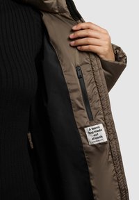 Pruun polsterdatud jakk musta voodriga, lukuga tasku ja kangast sildiga, millel on kiri "Soe kallistus tehtud kangast." Tekstuuriga välimine kiht.