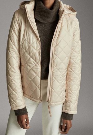 Quilted cream hooded jacket worn over a brown turtleneck sweater and white pants, shown from neck to hips.