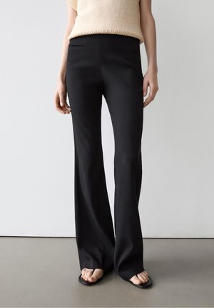 Person trägt schwarze, hoch taillierte Schlaghosen und ein beiges ärmelloses Strickoberteil, steht barfuß auf einem grauen Boden vor einer weißen Wand.