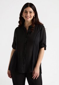 Camisa negra de botones con cuello, mangas cortas enrolladas y aberturas laterales. Fabricada en una tela ligera y suave. Detalles mínimos.