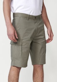 Uomo in pantaloni cargo verdi oliva con molte tasche e una maglietta bianca, in piedi davanti a uno sfondo neutro.