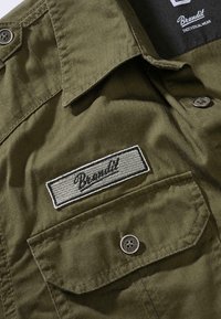 Olivengrønn skjorte med knapper, som har en brystlomme, brodert "Brandit"-merke og en stoffstruktur med tydelige sømdetaljer.