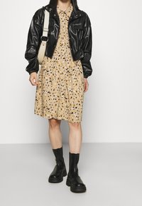 Veste noire en cuir courte, robe beige avec des motifs abstraits noirs et bruns, et bottines à semelle épaisse noires. Sac bandoulière visible.