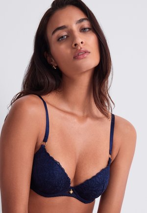 Jeune femme aux longs cheveux foncés portant un soutien-gorge en dentelle bleu marine à fines bretelles sur un fond clair uni.