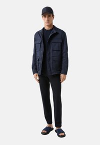 Marineblaues Jacket mit mehreren Fronttaschen, über einem marineblauen Oberteil getragen, kombiniert mit schwarzen Hosen und dunklen Sandalen, alles auf einem einfarbigen Hintergrund.