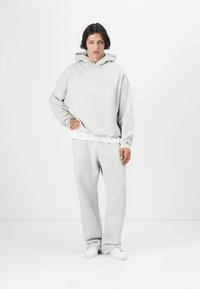 Lys grå hettegenser med stor hette, løs passform og ribbestrikkede mansjetter, kombinert med matchende baggy bukser og hvite joggesko. Jevn stofftekstur.