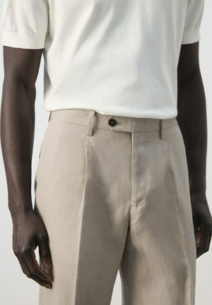 Man wearing beige high-waist trousers with pleats and a black button, paired with a short-sleeve white knit top.
