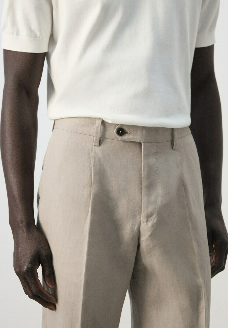 Homme portant un pantalon beige taille haute avec des plis et un bouton noir, assorti à un haut en maille blanc à manches courtes.