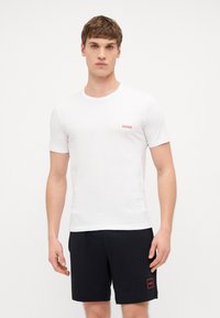 White cotton t-shirt with short sleeves, round neck, and red "HUGO" logo on the left chest. Paired with black shorts featuring a red logo.