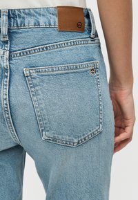 Κοντινό πλάνο της πίσω τσέπης ενός τζιν παντελονιού σε ανοιχτό μπλε χρώμα, με αντιθετική ραφή, δερμάτινο ταμπελάκι και μικρό κυκλικό λογότυπο κοντά στην άκρη της τσέπης.