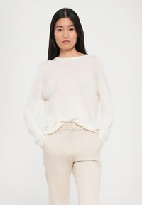 Pull en tricot léger de couleur crème, à manches longues et à encolure ronde, associé à un pantalon beige ajusté. Texture douce, coupe décontractée.