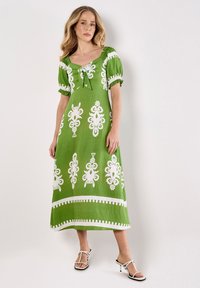 Grünes Leinenkleid mit weißen Blumendrucken, quadratischem Ausschnitt, Puffärmeln, Bindeband vorne und einem knielangen Saum. Weiße Sandalen.