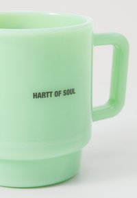 Taza verde translúcida con un cuerpo redondeado, asa gruesa y una impresión que dice "HARTT OF SOUL" en letras negras negritas cerca de la base.