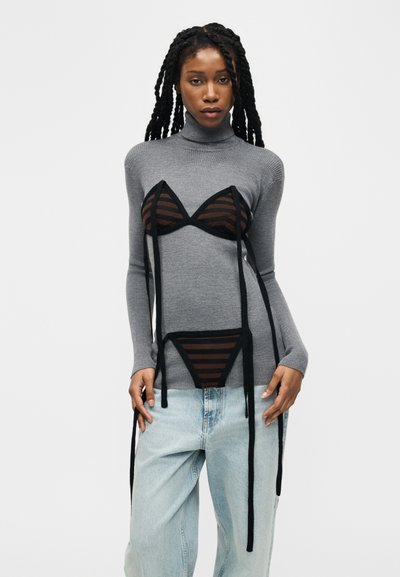 Femme aux longs cheveux tressés portant un pull à col roulé gris superposé à une lingerie noire rayée et un jean bleu clair, sur fond blanc.