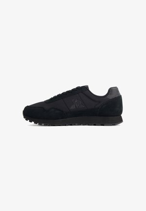 Black low-top sneaker with suede and fabric panels, lace-up closure, ridged sole, and subtle embossed logo on the side.
