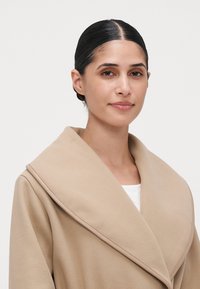 Kvinne med mørkt hår bundet bak, iført en beige frakk med bred krage over en hvit topp, som ser litt smilende ut mot en ensfarget bakgrunn.