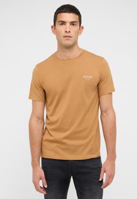 T-shirt à manches courtes et col rond en coton marron clair avec un petit logo blanc sur la poitrine, porté avec un jean gris foncé.