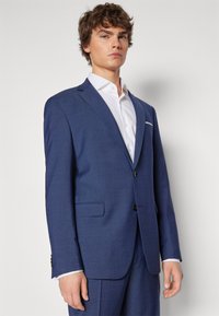 Blau, strukturiertes Sakko mit zwei Knöpfen, Reverskragen und Taschen. Kombiniert mit einem weißen Hemd mit Kragen aus glattem Stoff.