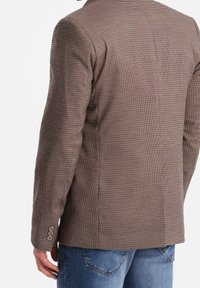 Brązowa marynarka w kratę z teksturowanego materiału; charakteryzuje się dopasowanym fasonem, pojedynczym rozcięciem z tyłu i mankietami na trzy guziki. Noszona na niebieskich dżinsach.