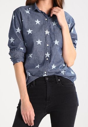 Kvinde iført en blå skjorte med knapper og hvide stjerner og sorte jeans, med den ene hånd, der rører ved hendes hals og den anden hvilende langs siden.