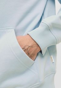 Sudadera azul claro con un bolsillo tipo canguro, puños acanalados y tela suave. Una mano con un tatuaje está colocada en el bolsillo.