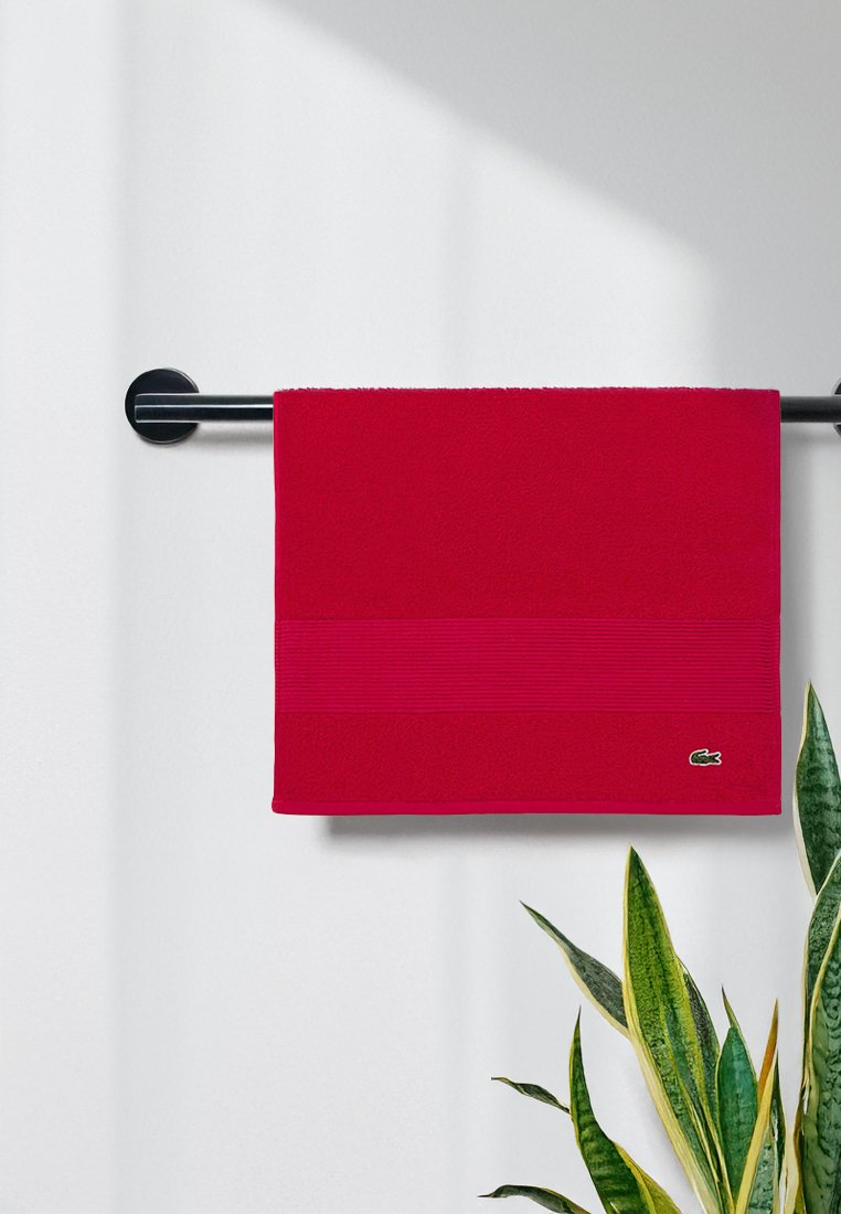 Toalla de algodón roja con banda de acento horizontal texturizada, exhibida en un toallero negro, junto a una planta verde. Logo de Lacoste en la esquina.