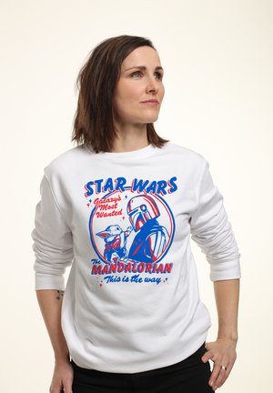 Kobieta w białej bluzie z czerwoną i niebieską grafiką Star Wars przedstawiającą Mandalorianina i Grogu z tekstem "Galaxy's Most Wanted" oraz "This is the way."