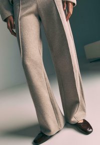 Béžové nohavice so širokými nohavicami vyrobené z mäkkej tkaniny, s hladkou textúrou, bočnými švami a jemným rozšírením na leme. V páre s tmavohnedými topánkami.