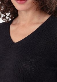 Haut noir en maille avec un col en V, présentant un motif texturé et une coupe ajustée. Le tissu semble léger et légèrement extensible.