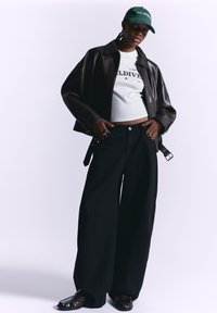 Sorte brede jeans, fitted hvid top og en mørk læderjakke. Grøn kasket med tekst. Skinnende mørke sko fuldender looket.
