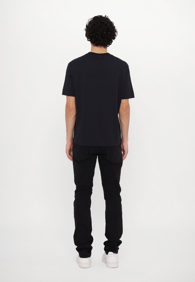 Tricou negru cu mânecă scurtă, croială lejeră, țesătură netedă. Asortat cu blugi negri strâmți și adidași albi. Fără detalii de design notabile vizibile.