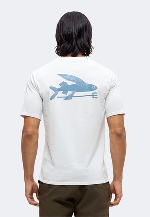 Mand med skulderlangt mørkt hår iført en hvid t-shirt med en blå flyvende fisk grafik på ryggen.