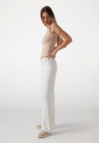 Fitted beige tank top with white wide-leg jeans. Jeans feature front pockets and gold-tone hardware. Model stands sideways, showing outfit fit.
