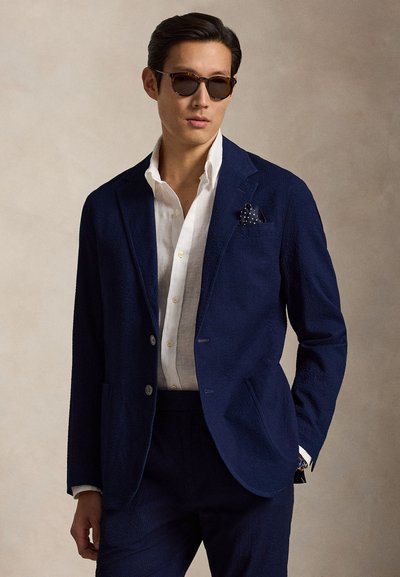 Άνδρας με σκούρο μπλε κοστούμι, λευκό πουκάμισο, μαντηλάκι με πουά στην τσέπη και γυαλιά ηλίου, στέκεται μπροστά σε μια μονόχρωμη μπεζ φόντο.