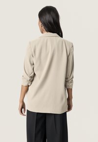 Blazer beige con maniche arrotolate, tessuto morbido, colletto sagomato e taglio dritto, abbinato a pantaloni neri a gamba larga.