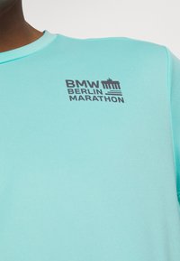 Tyrkysová krátká rukávová košile s kulatým výstřihem, na které je tištěné logo "BMW BERLIN MARATHON" v tmavě šedé barvě. Hladká textura, atletický střih.