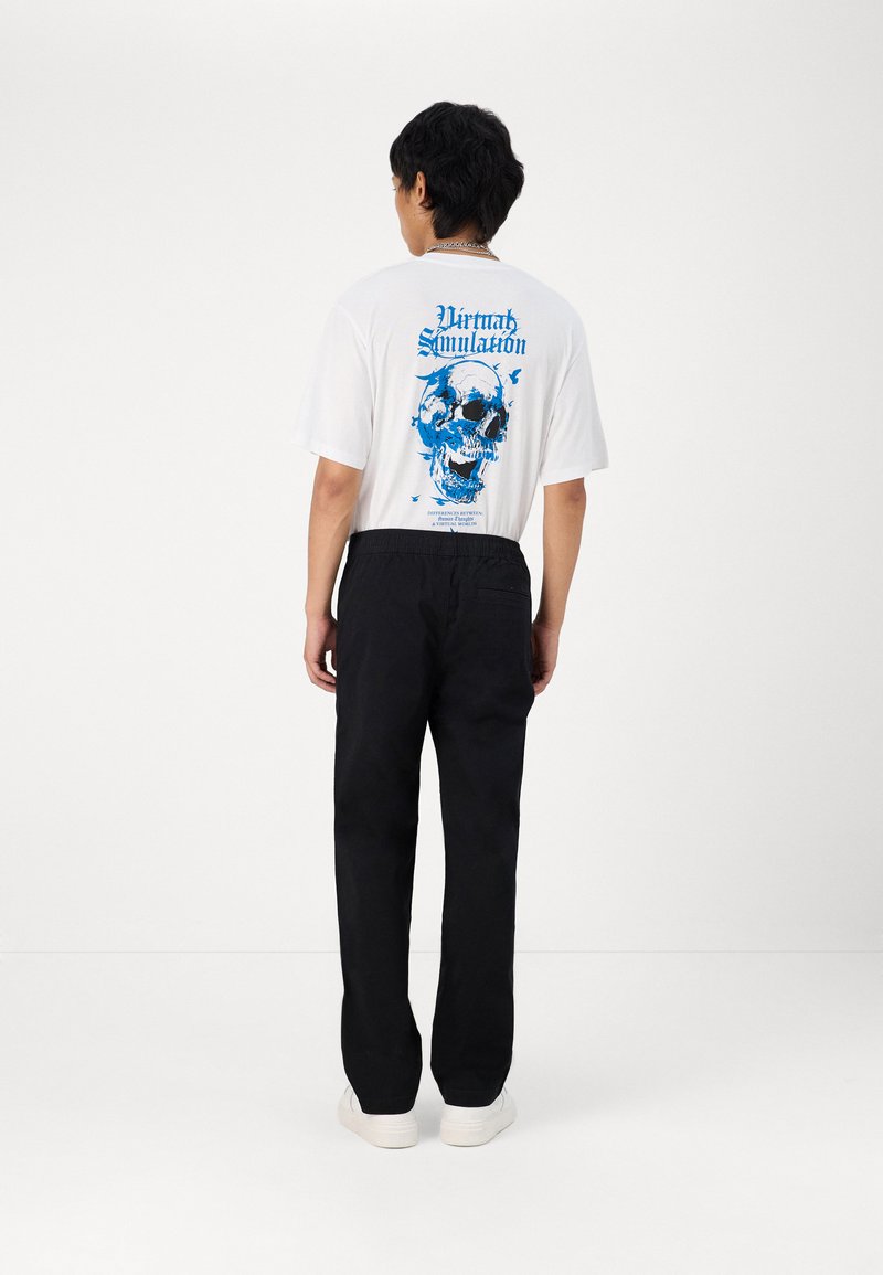 Weißes T-Shirt mit blauem grafischen Totenkopfmuster auf dem Rücken, kombiniert mit schwarzen Hosen und weißen Turnschuhen. Schlichte und lässige Passform.