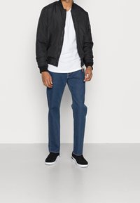 Hombre vistiendo una chaqueta bomber negra, camiseta blanca, jeans azules y zapatillas negras, de pie sobre un suelo gris liso con fondo blanco.