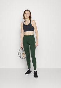 Top desportivo preto, leggings verdes de cintura alta com detalhes nas costuras, sapatilhas desportivas pretas, segurando uma corda de saltar, fundo minimalista.