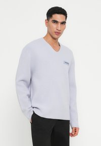 Gai pelēks, adīts pulovers ar V-veida kaklu, garām piedurknēm un mazu zilu logo plāksteri uz krūtīm. Mīksta tekstūra.