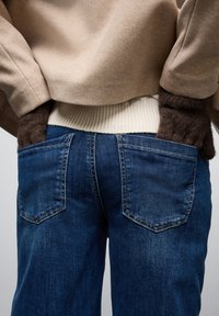 Jeans in denim blu con un design aderente, caratterizzati da due tasche posteriori e dettagli di cucitura. Abbinati a un top beige e testurizzato con polsini marrone scuro.